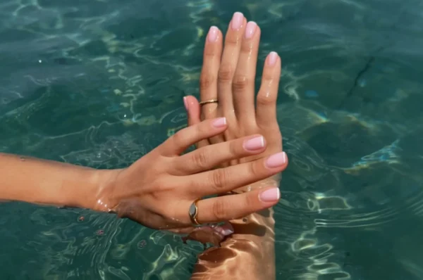 Fotografija prikazuje nježne ženske ruke s elegantnom, prirodnom manikurom u nježno ružičastoj nijansi. Slika prikazuje kvalitentnu njegu noktiju ljeti.