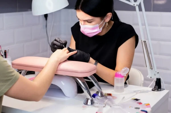 Nail tehničarka s maskom na licu i crnim rukavicama radi manikuru klijentici u profesionalno uređenom salonu s bijelim stolom i lampom za nokte.