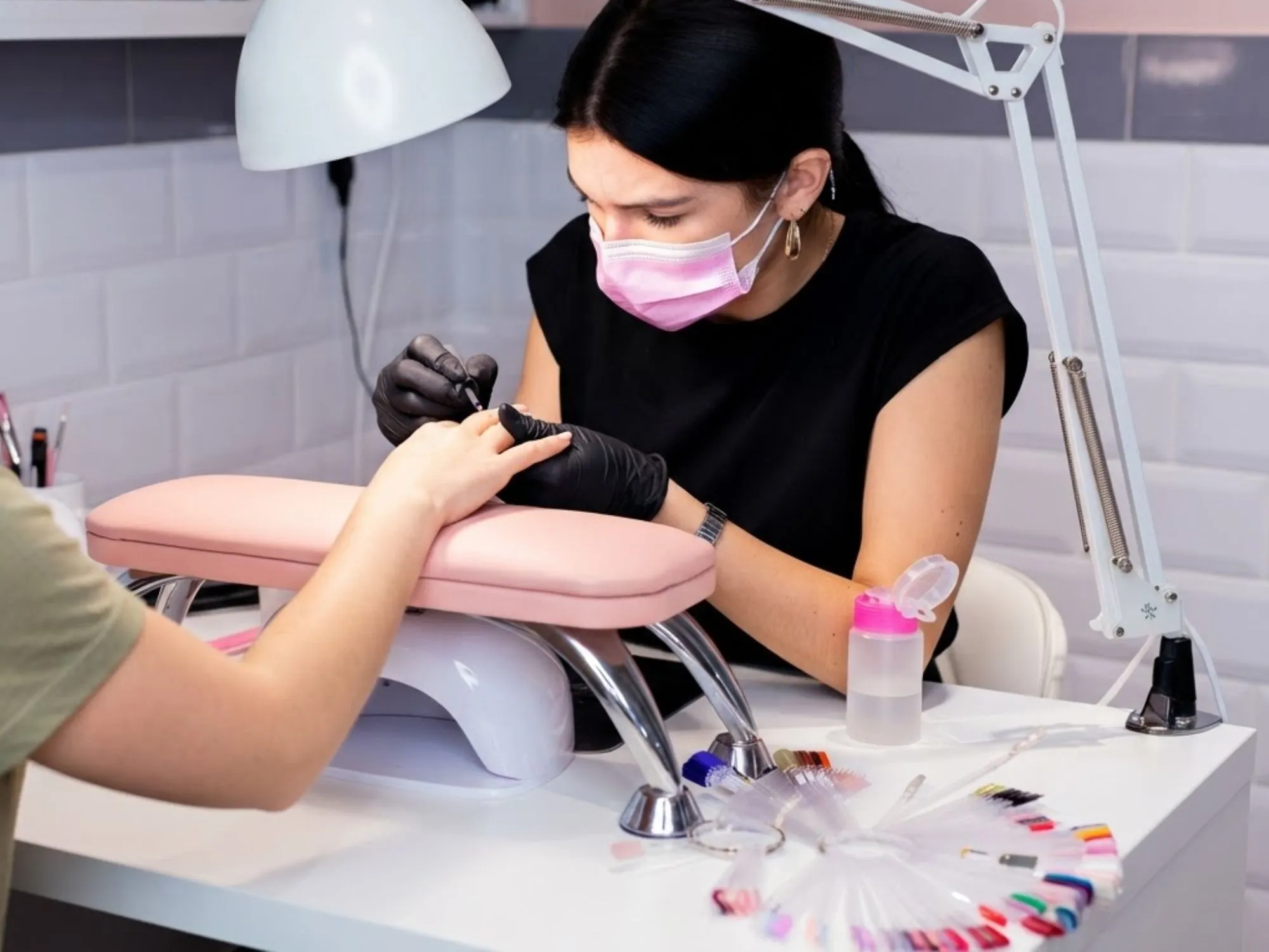 Nail tehničarka s maskom na licu i crnim rukavicama radi manikuru klijentici u profesionalno uređenom salonu s bijelim stolom i lampom za nokte.